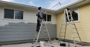 A homeowner using a roller on a ladder while learning how to paint a house exterior.